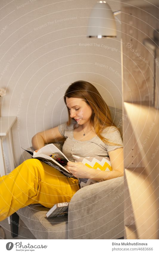 Young beautiful woman wearing gray and yellow sweatpants reading a book at home, sitting in cozy chair. education and knowledge. Literature and fiction books.  Cozy home room.