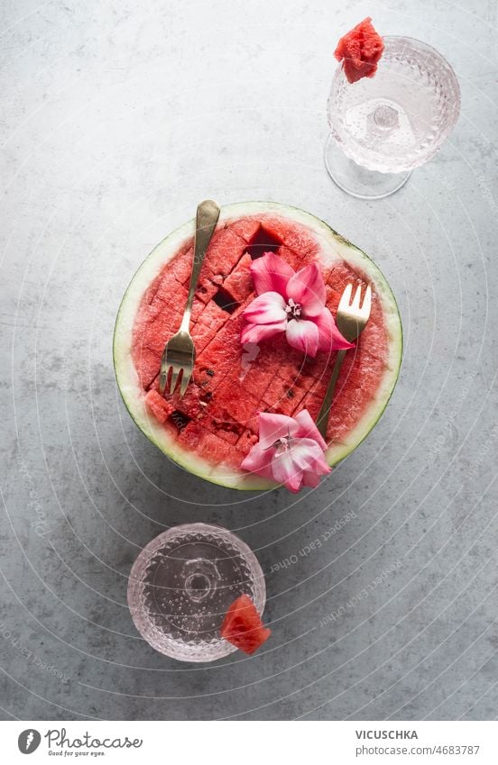 Halved watermelon with golden forks and flowers at grey concrete kitchen table with champagne glasses halved summer food drink concept refreshing fruit cocktail
