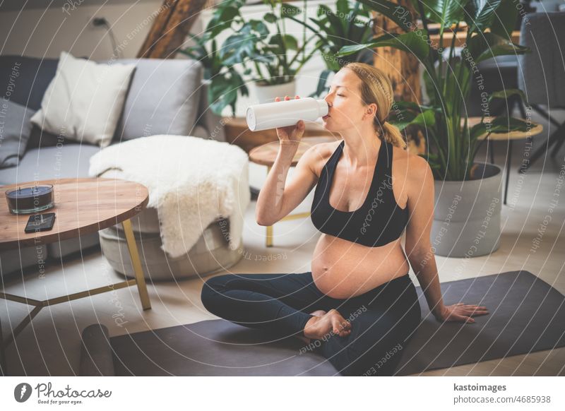Young happy and cheerful beautiful pregnant woman taking a break, hydrating, drinking water from the botle after home well being workout progrem mother yoga