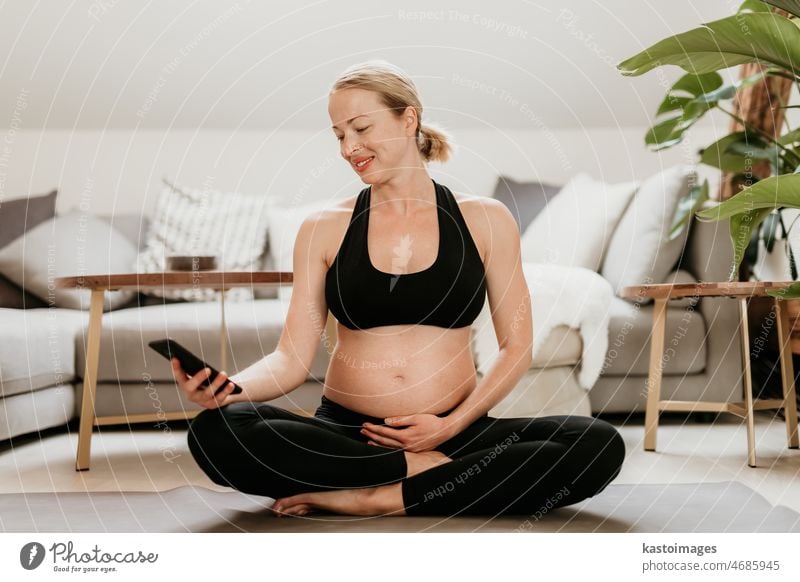 Pregnancy and technoloy supported healthy lifestyle concept. Cheerful happy  pregnant woman using smart phone application while exercising on yoga mat  on living room floor at home. - a Royalty Free Stock Photo