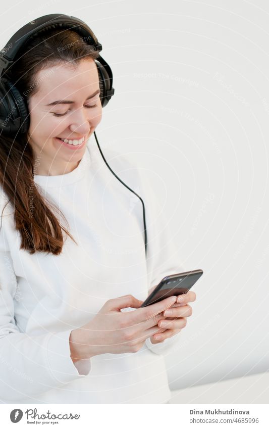 Young brunette millennial woman using mobile phone and listening to music in headphones, smiling.  Young adult in white sweatshirt using technology isolated on white background.