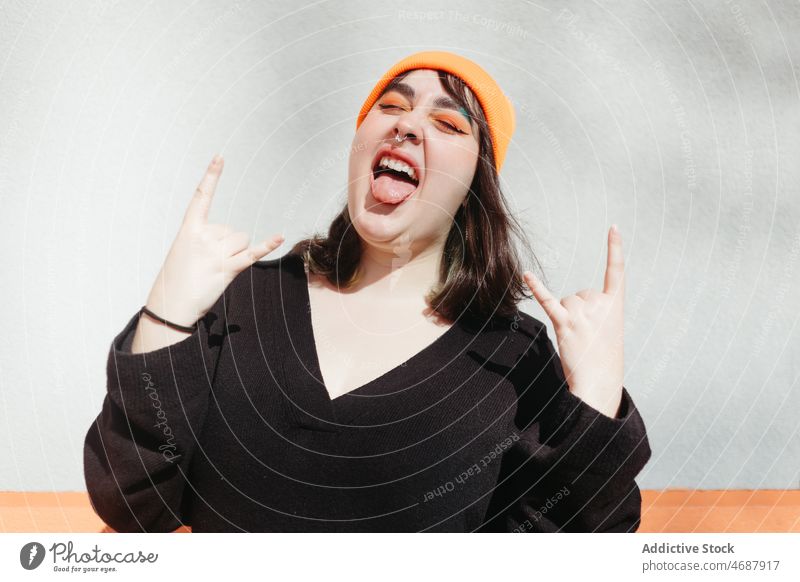 Woman showing rock and roll gesture woman style horn show tongue rebel street city informal trendy design wall building lady cheerful young female content