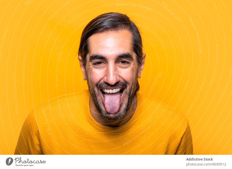 Cheerful man showing tongue in studio show tongue grimace make face carefree funny appearance silly mood playful beard portrait male tongue out yellow unshaven