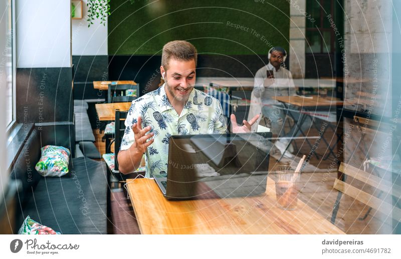 Young man with earphones and laptop in a coffee shop happy surprised success win casual wear free wifi smiling working through the glass beard reflection young