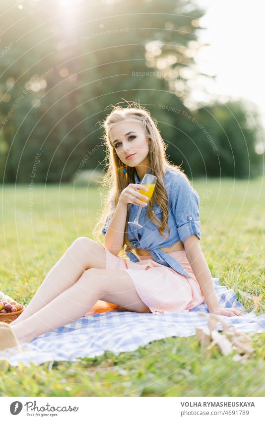Portrait of a young woman holding orange juice in a glass in an open park. person blanket sitting happy picnic book outside smile happiness pretty female hat