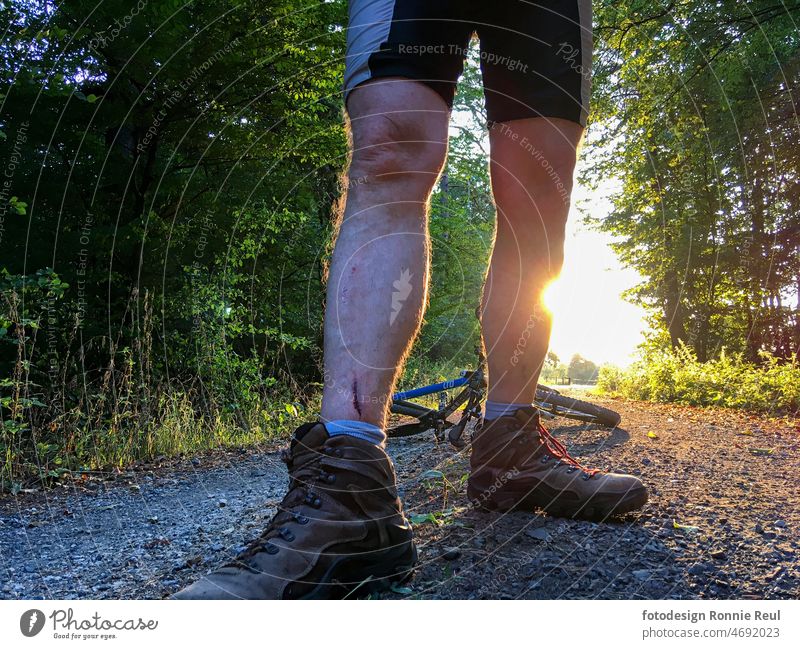 Wounded mountain biker backlit on a gravel forest trail. Mountain bike Bicycle Accident Sudden fall Legs Risk Pain Forest forest path trees Sun Back-light