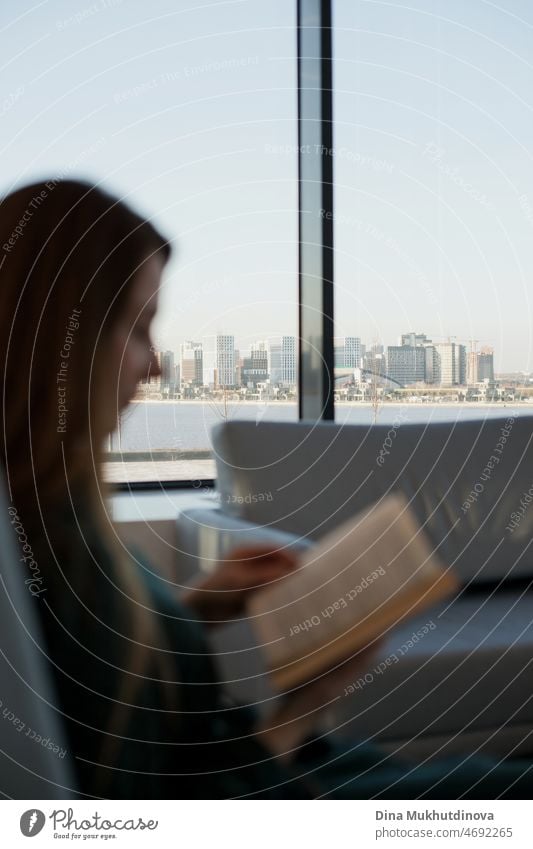 Woman reading a book sitting by the window with a cityscape view in it on a sunny day. Modern casual lifestyle - blurred silhouette of woman with a book and city landscape view in focus. Reading and education.