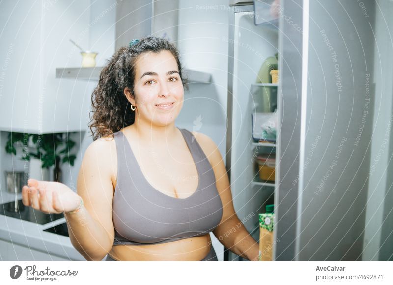 Curly woman doubting about what to eat from the fridge at the kitchen home to lose weight. Training clothes. Getting fit for the summer concept.Beach body preparation, healthy life