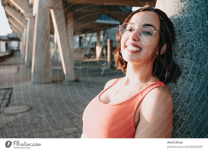 Young african Moroccan girl in training clothes with glasses smiling to camera while resting against a wall. Sports on people with vision problems concept. Urban place with copy space during sunset