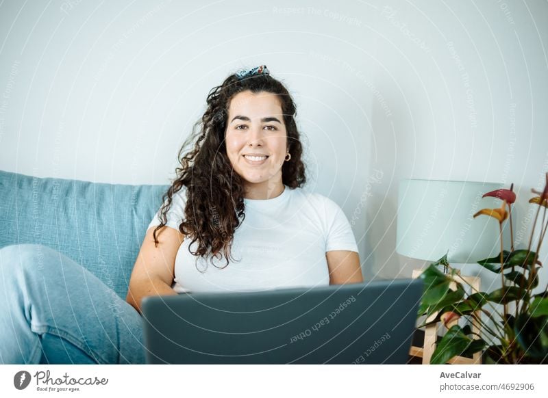 Happy plus size curly young woman smiling to camera while using a laptop to prepare,and do work online at home, remote tasking. Modern city flat. Getting the bills paid. Entrepreneur freelancer women
