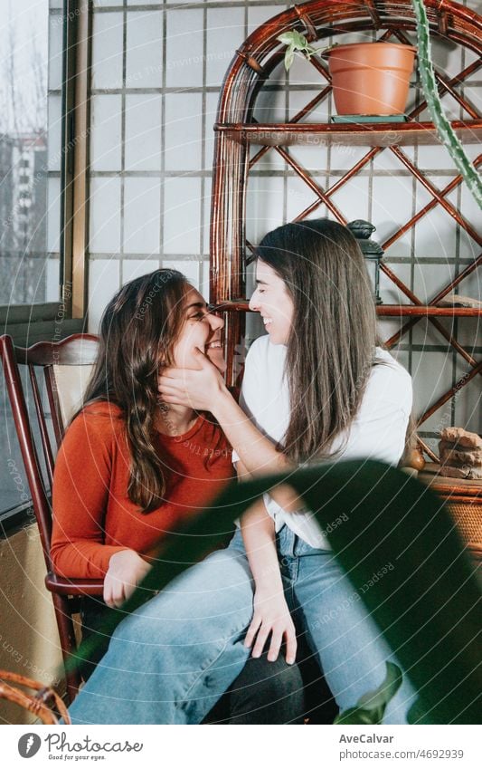 Young and modern lesbian couple cuddling and smiling to each other while sitting on a modern chair on a gallery on a flat. Happy together. LGBTI+ love. Pride and right fighting concept