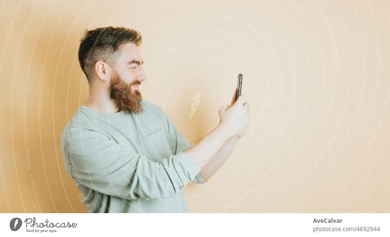 Young man bearded hipster style holding a mobile phone and taking a photo, yellow background. Emotion about receiving a good new, removable background with copy space. Young people social network