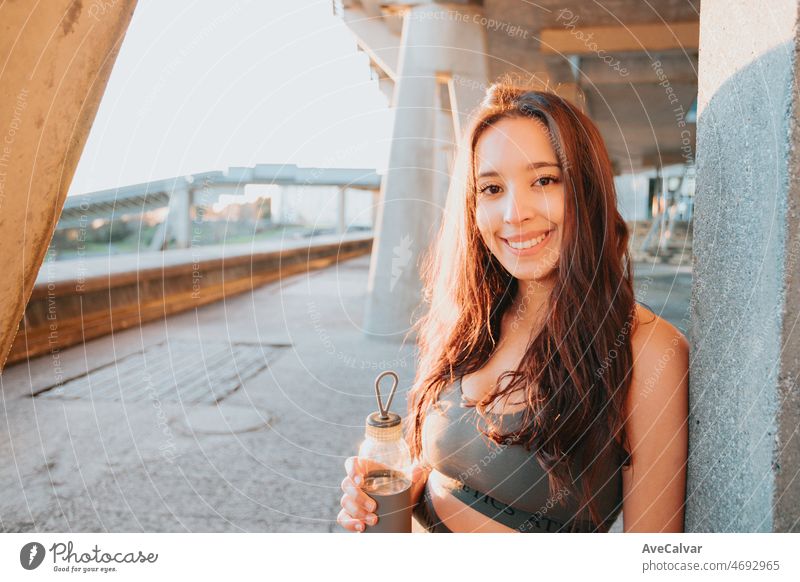Young girl in training clothes with long hair holding water smiling to camera while resting against a wall. Sports on people with vision problems concept. Urban place with copy space during sunset
