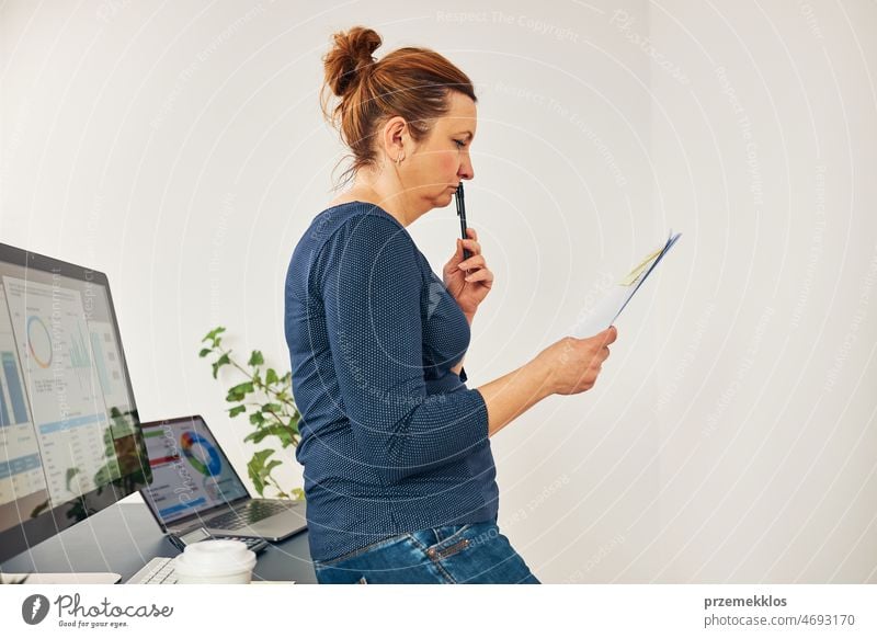 Woman entrepreneur focused on solving difficult work. Confused businesswoman thinking hard holding doccuments standing at her desk in office frustration problem