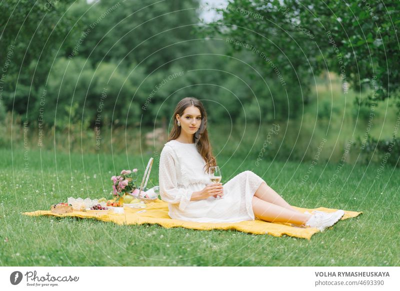 A beautiful young woman in a white dress drinks wine in the garden on a picnic. Summer vacation in solitude basket bottle plaid baguette healthy nature book