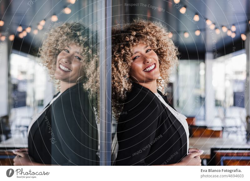 portrait of beautiful smiling business woman leaning on glass in cafe. business concept afro hispanic mobile phone city skyscraper building young curly hair