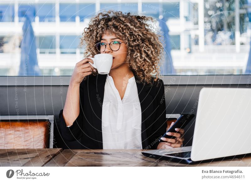 beautiful hispanic afro business woman wearing eye glasses in cafe working on laptop, mobile phone computer networking drinking coffee terrace outdoors goggles