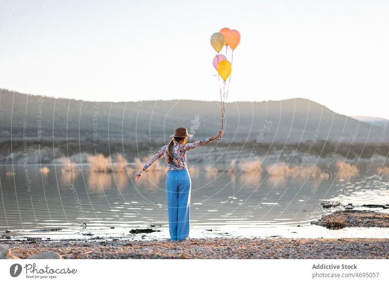 Positive woman with balloons near lake shore water countryside embankment coast nature style female hat colorful waterside arms raised summer bright river