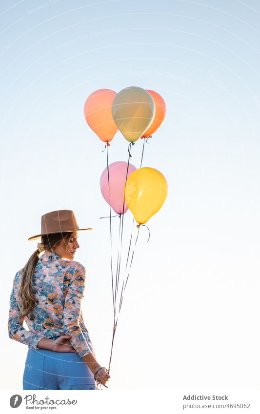 Anonymous woman with balloons against clear sky countryside nature style female hat colorful summer bright outfit waterfront range leisure evening mountain