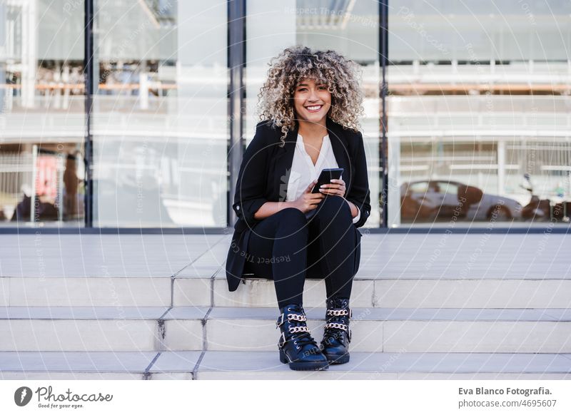 beautiful smiling business woman using mobile phone sitting on stairs in city. Buildings background afro hispanic skyscraper building young curly hair finances