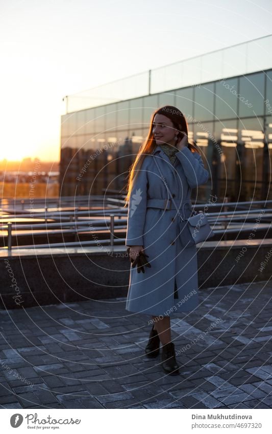 Woman in stylish blue coat leaving office at sunset. Beautiful attractive young woman walking on the street at sunset or sunrise early in the morning near modern architecture buildings in the city. Casual candid lifestyle. Full body portrait.