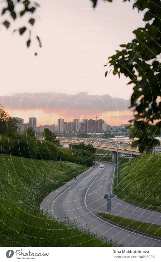 Cityscape with an empty road and buildings at sunset and green grass and tree leaves. Summer in the city vertical background. Warm summer nights urban background.