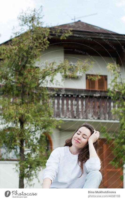 Young woman in white sweatshirt and light blue jeans sitting near a house on green grass in summer or spring. Casual lifestyle portrait. beautiful natural