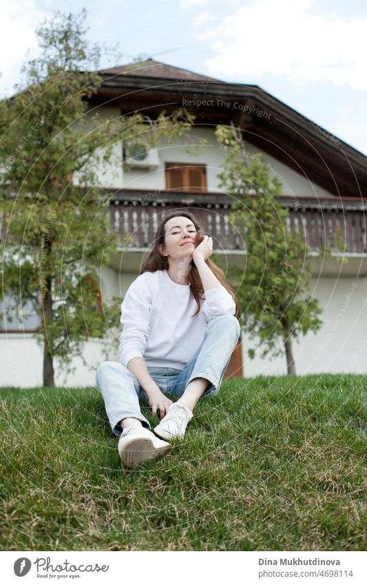 Young woman in white sweatshirt and light blue jeans sitting near a house on green grass in summer or spring. Casual lifestyle full body portrait. beautiful