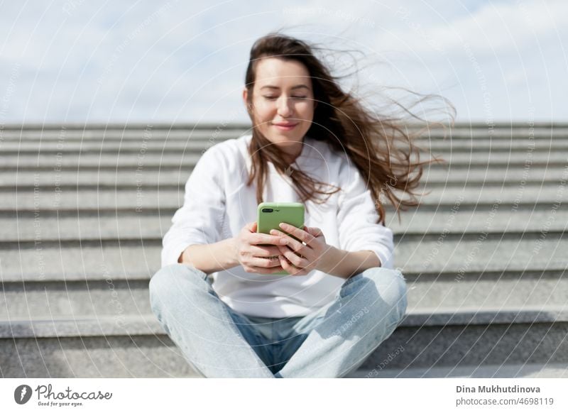 Young millennial woman in white shirt taking a selfie or videochatting on mobile phone sitting on the steps of a building. Female student using technology sitting on staircase steps of university campus. Real people with mobile phones.