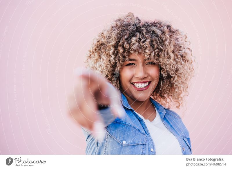 beautiful happy hispanic woman afro hair outdoors in spring pointing with hands. pink background touch smiling city casual clothing lifestyle curly hair urban