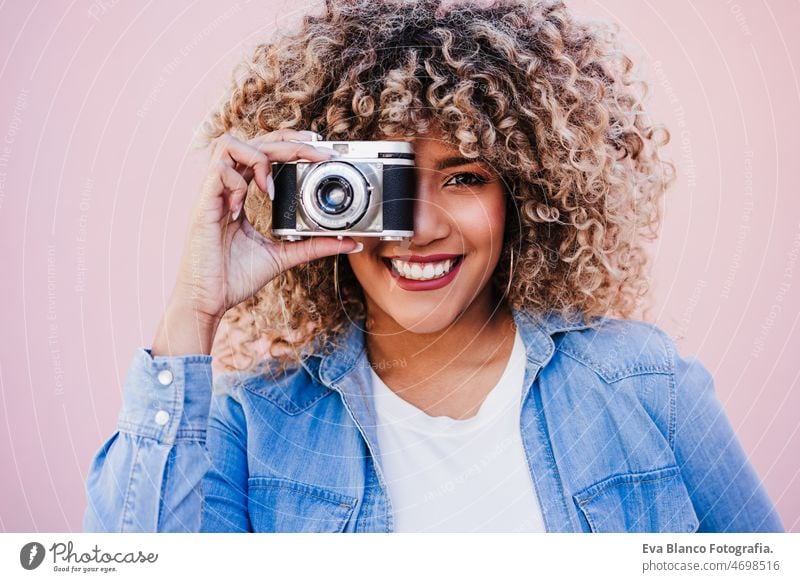 beautiful happy hispanic woman with afro hair holding vintage camera. pink background photographer city touch smiling spring casual clothing lifestyle