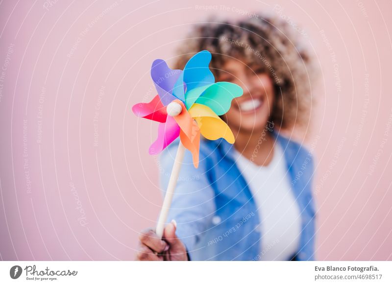 beautiful happy hispanic woman with afro hair holding colorful pinwheel. pink background,wind energy windmill sustainability reduce reused recycle city smiling
