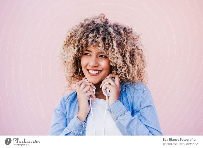 portrait of smiling hispanic woman with afro hair in city using mobile phone and headset. lifestyle selfie technology internet wireless picture device music