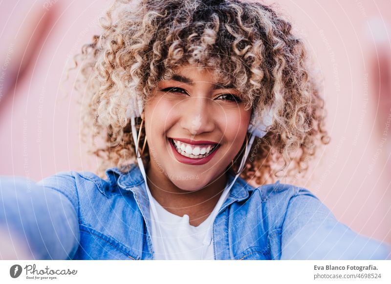 portrait of smiling hispanic woman with afro hair in city using mobile phone and headset. lifestyle selfie technology internet wireless picture device music