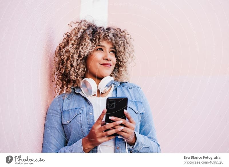 portrait of smiling hispanic woman with afro hair in city using mobile phone and headset. lifestyle music listening spring pink young people african street girl