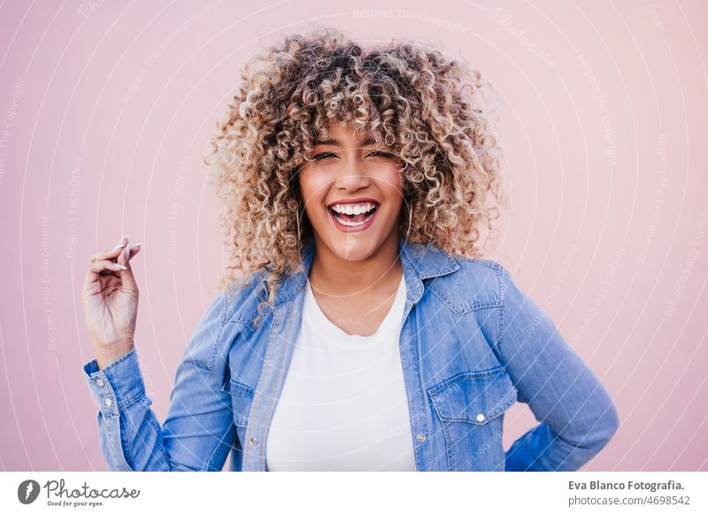 portrait of smiling hispanic woman with afro hair in city during spring. Urban lifestyle happy pink casual clothing curly hair outdoors urban adult laughing