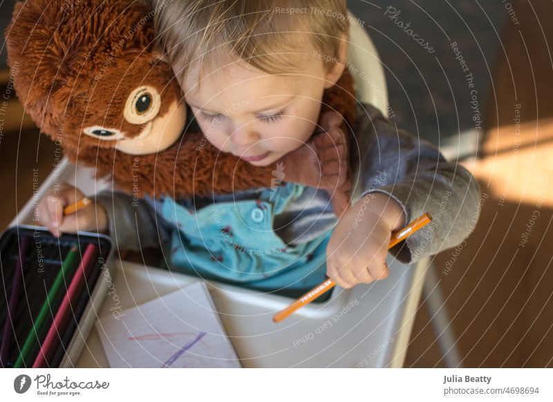 Toddler girl drawing with a colored pencil while seated in high chair with a stuffed chimpanzee toy child education school childhood kid paper pre-writing art