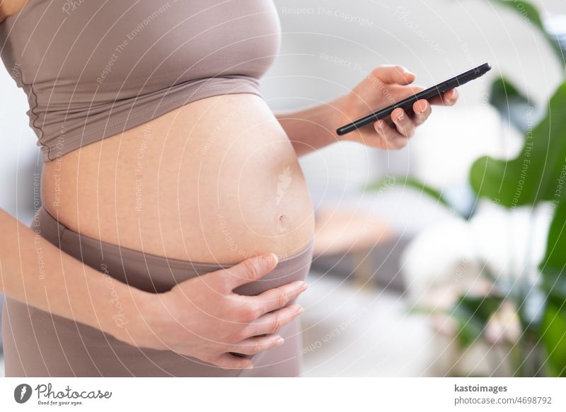 Close-up photo of pregnant female belly. Woman holding and using mobile smart phone application at home interiors. Pregnancy, technology, online shopping, preparation and expectation concept.