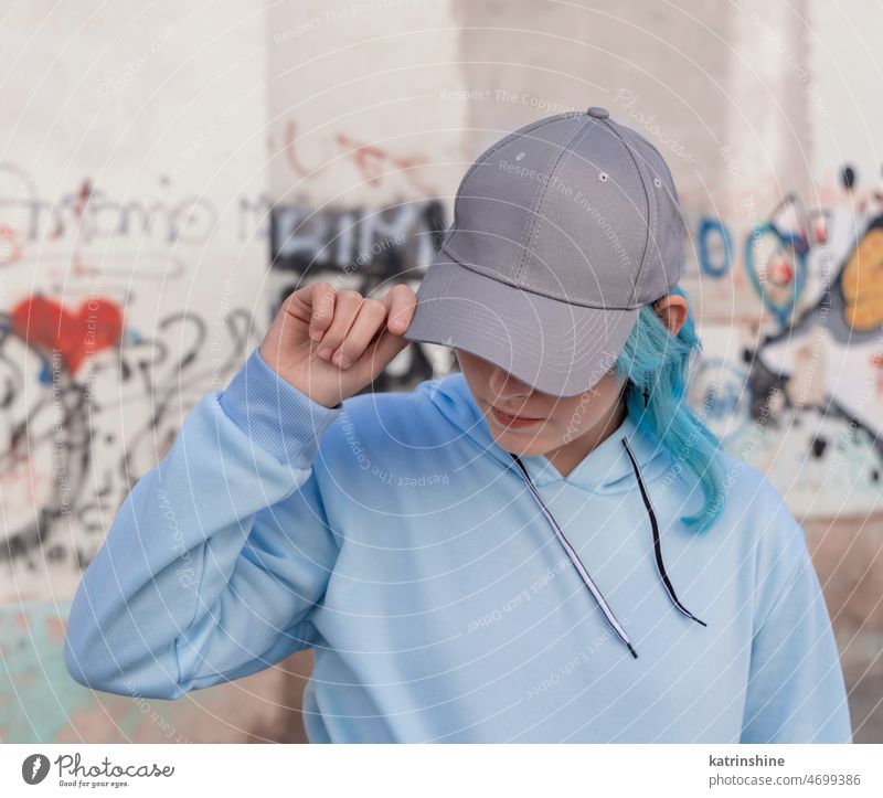 Blue haired Teenage girl in blue hoodie and baseball cap staying against graffiti wall Teenager light blue mockup blue haired teen girl outdoors urban modern