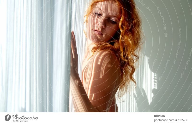 Portrait of a beautiful young attractive sexy redhead woman, partially obscured by the white threads of a string curtain, that conjures up a shadow play on the face, copy space
