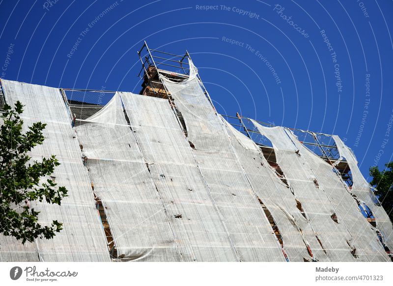 Scaffolding with tattered safety net during the renovation of the facade of a large old building in front of a blue sky in sunshine in the district of Sachsenhausen in Frankfurt am Main in the German state of Hesse