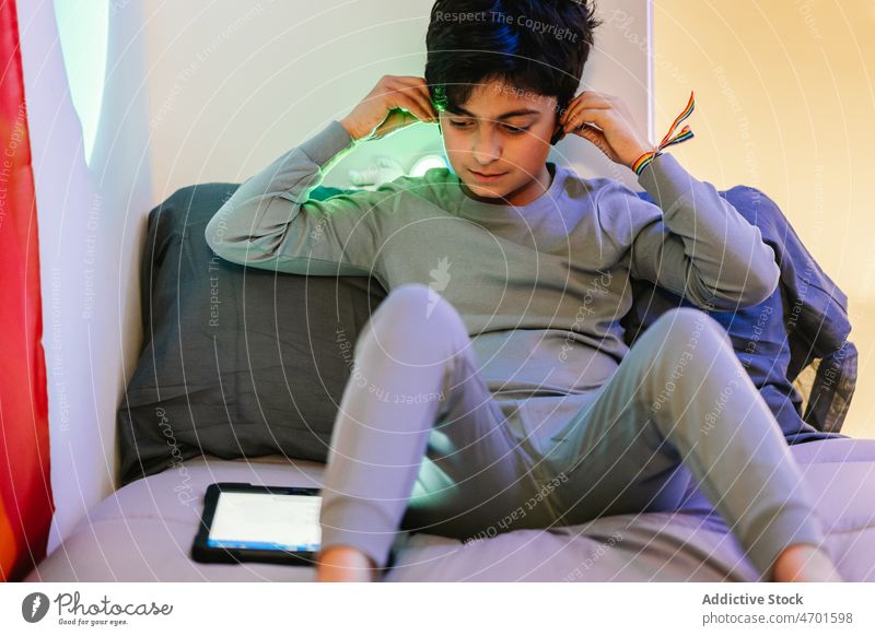 Ethnic teen boy putting on earbuds and using tablet watch bed non binary bracelet flag wireless rainbow minority gadget earphones online device sit adjust