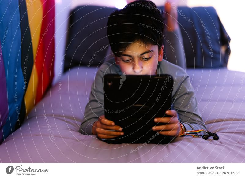 Ethnic boy using tablet on bed during bedtime at night watch bed time online rainbow flag lgbtq non binary browsing internet lying surfing kid connection gadget
