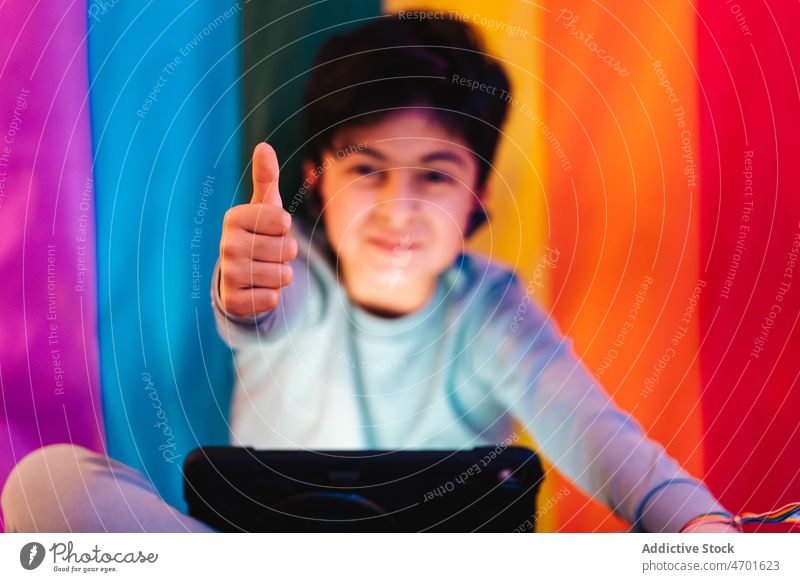 Ethnic boy in earbuds using tablet and showing thumb up gesture like recommend flag rainbow respect lgbtq minority gender solidarity equal tolerance watch