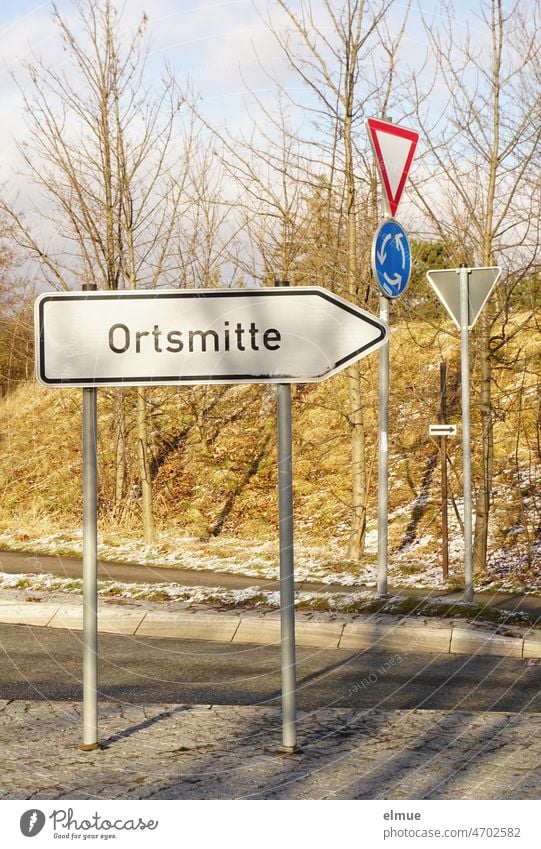 in the foreground on a traffic island, pointing to the right, the sign - Ortsmitte - and in the background the traffic signs - Kreisverkehr - and - Vorfahrt gewähren - in frosty sunny weather
