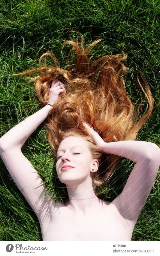 Portrait of a beautiful young sexy red-haired woman, raising her arms, lying in the spring sun relaxing on the green grass raises her arms Red hair Grass Girl