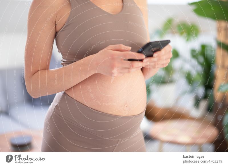 Close-up photo of pregnant female belly. Woman holding and using mobile smart phone application at home interiors. Pregnancy, technology, online shopping, preparation and expectation concept.