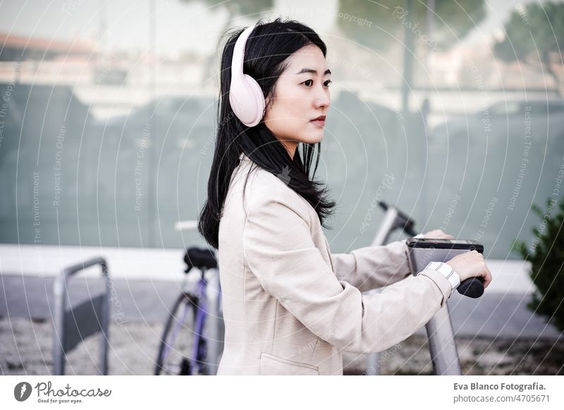 chinese business woman renting electric scooter in city,wearing headphones.sustainable transport music urban modern sustainability public transport asian young