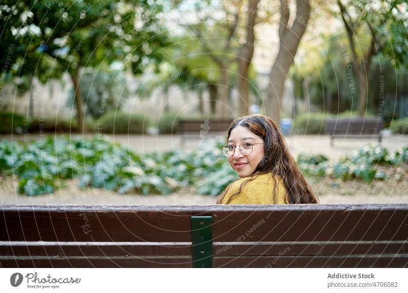 Positive woman sitting on bench park appearance feminine style rest plant leisure pastime young smile summer eyeglasses lady long hair youth female beautiful