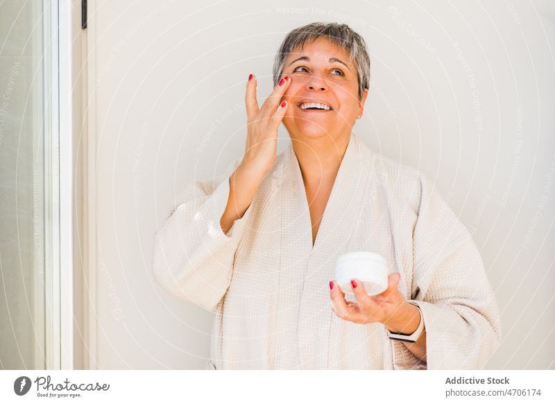 Cheerful elderly woman in bathrobe applying facial cream skin care moisture cosmetic smear treat body positive bathroom happy routine touch face smile adult